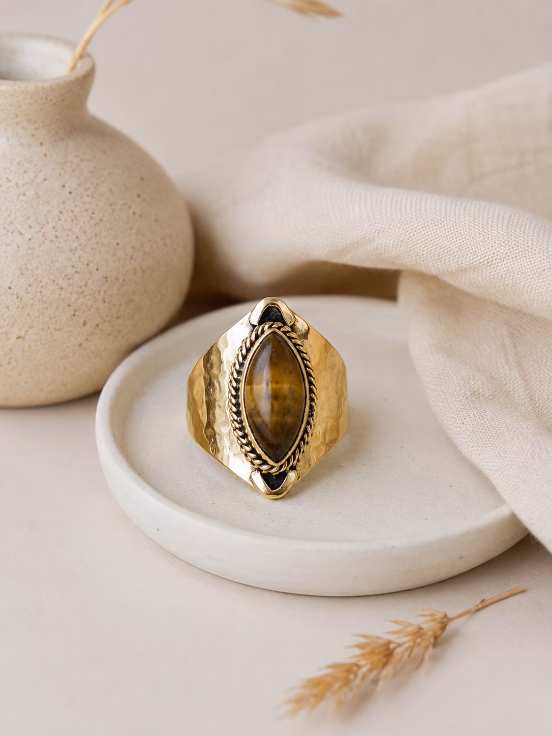 Bague-laiton-martele-sala-pierre-naturelle Bague en laiton martelé Salä avec pierre œil de tigre posée sur un support en céramique.