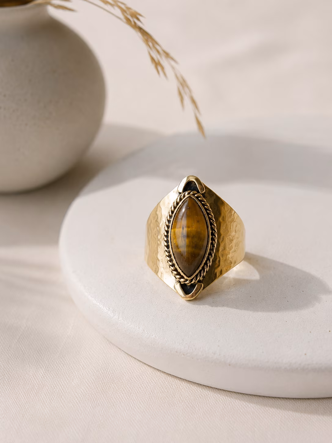 Bague-laiton-martele-sala-pierre-naturelle Bague en laiton martelé Salä avec pierre œil de tigre posée sur un support en céramique.